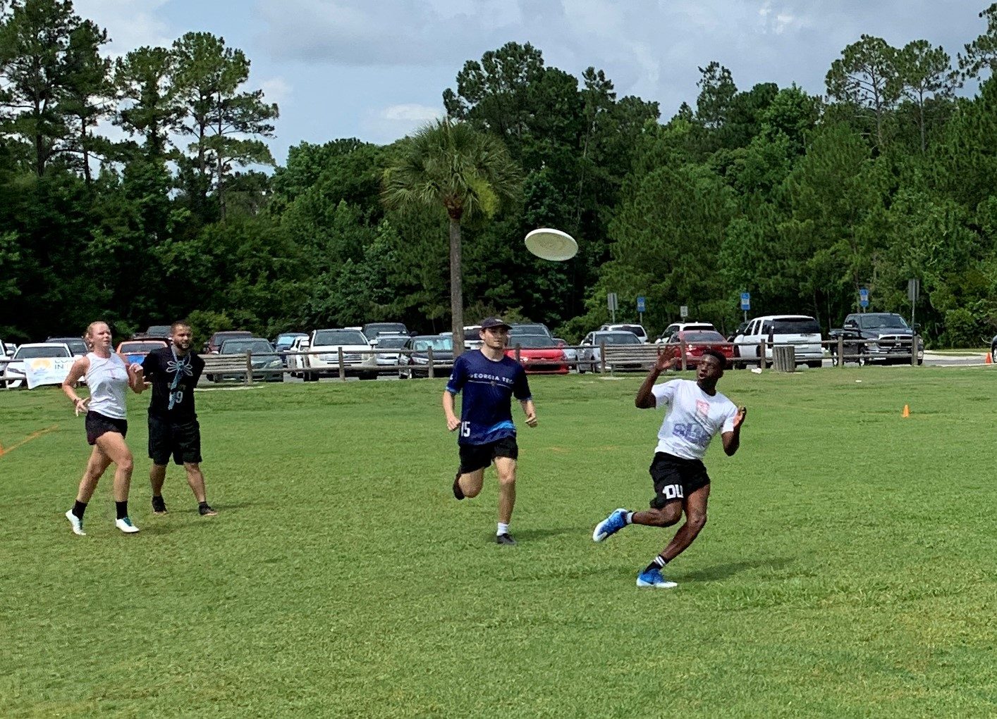 Group of competitors playing ultimate in an open field with cars in the background.