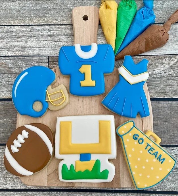 Football decorated cookie, Football goal post decorated cookie, yellow speaker with blue writing spelling "GO TEAM" with white polka dots cookie, blue cheerleader uniform with white v-shaped stripe cookie, blue football jersey with yellow "1" on center cookie, blue football helmet, all on small wooden cutting board with yellow, green, blue and brown icing bags next to it.