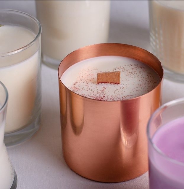 metallic rose colored candle frame with white candle inside with red specks and wooden wick surrounded by a purple candle and four white candles