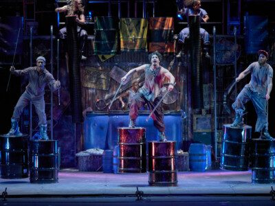 Stomp performers on stage in costume beating drum barrels