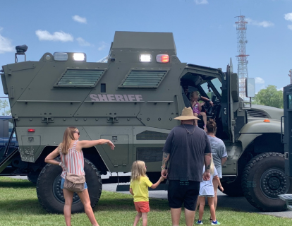 Family standing in front of a large green armored vehicle with "Sheriff" on the side.
