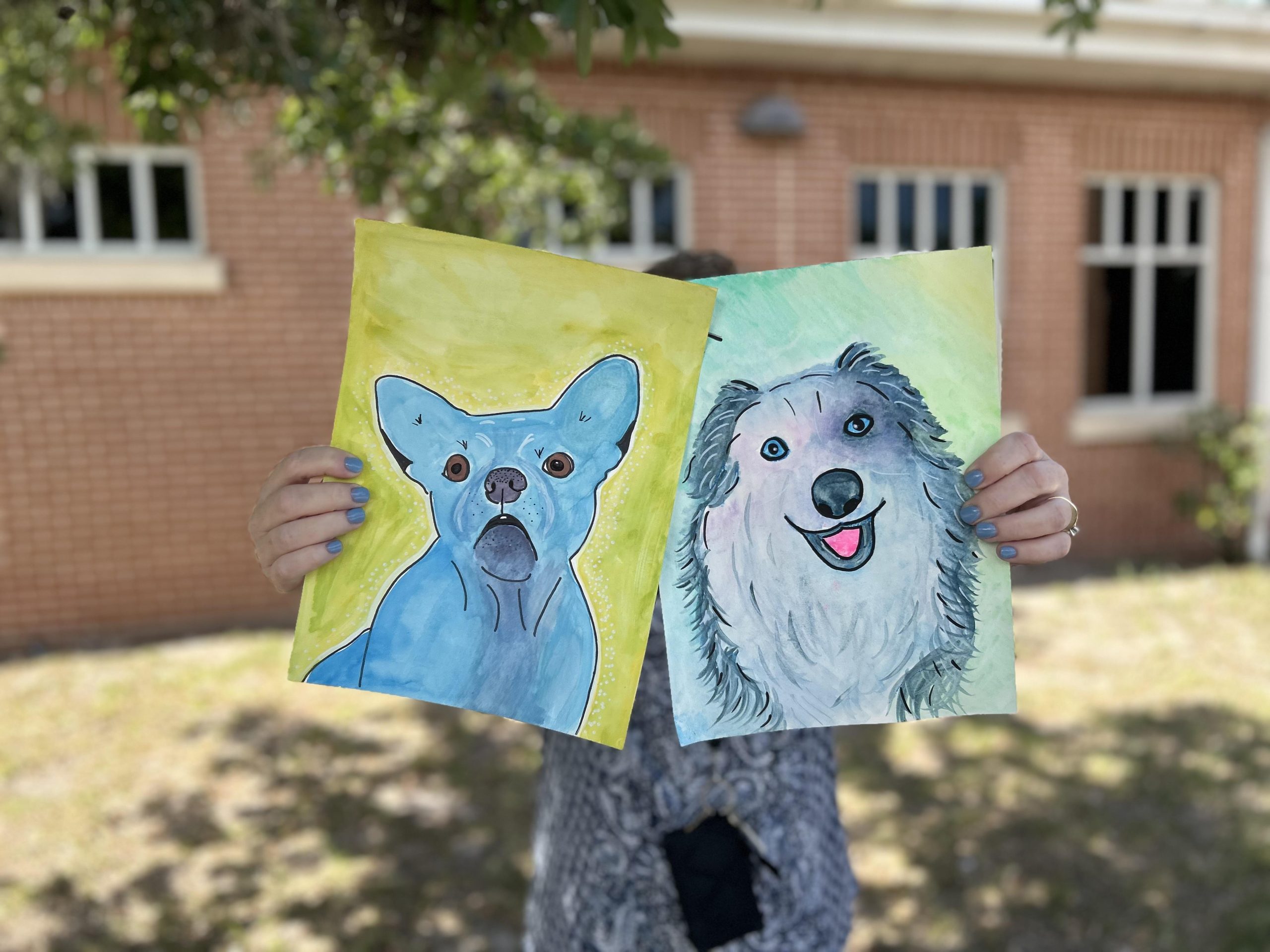 Pet Portraits Feature Photo Person holding two water color portraits of dogs.